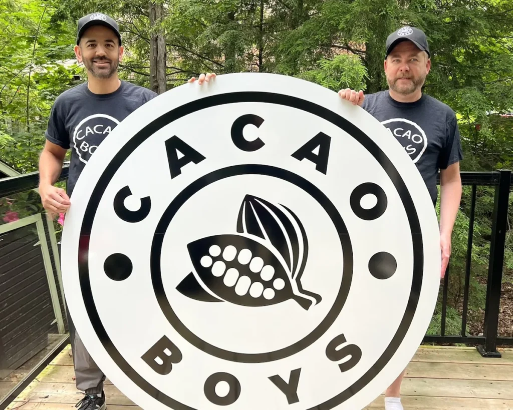 The Cacao Boys founders holding the Cacao Boys logo sign outside the Bala shop